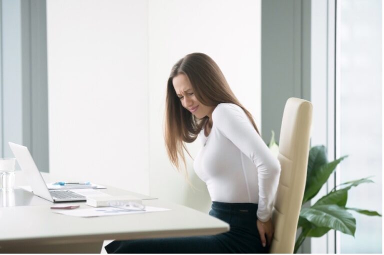 A woman sitting in the chair is in pain, suffering from hemorrhoids caused by untreated irritable bowel syndrome. She feels severe pain and experiences difficulty sitting